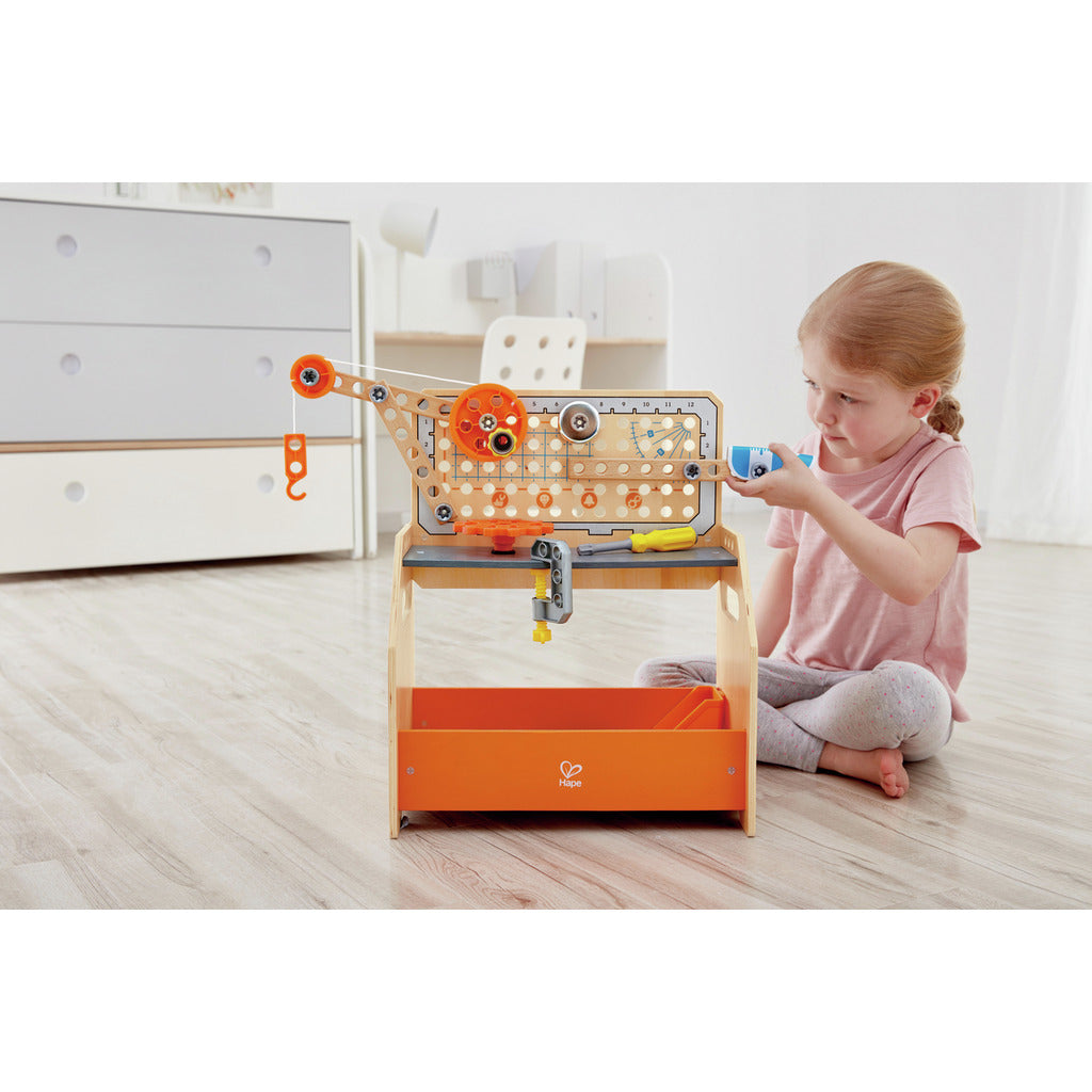 The wooden Hape Scientific Workbench, featuring a functional crane, gears, and an orange storage tray, is being used by a young girl sitting indoors