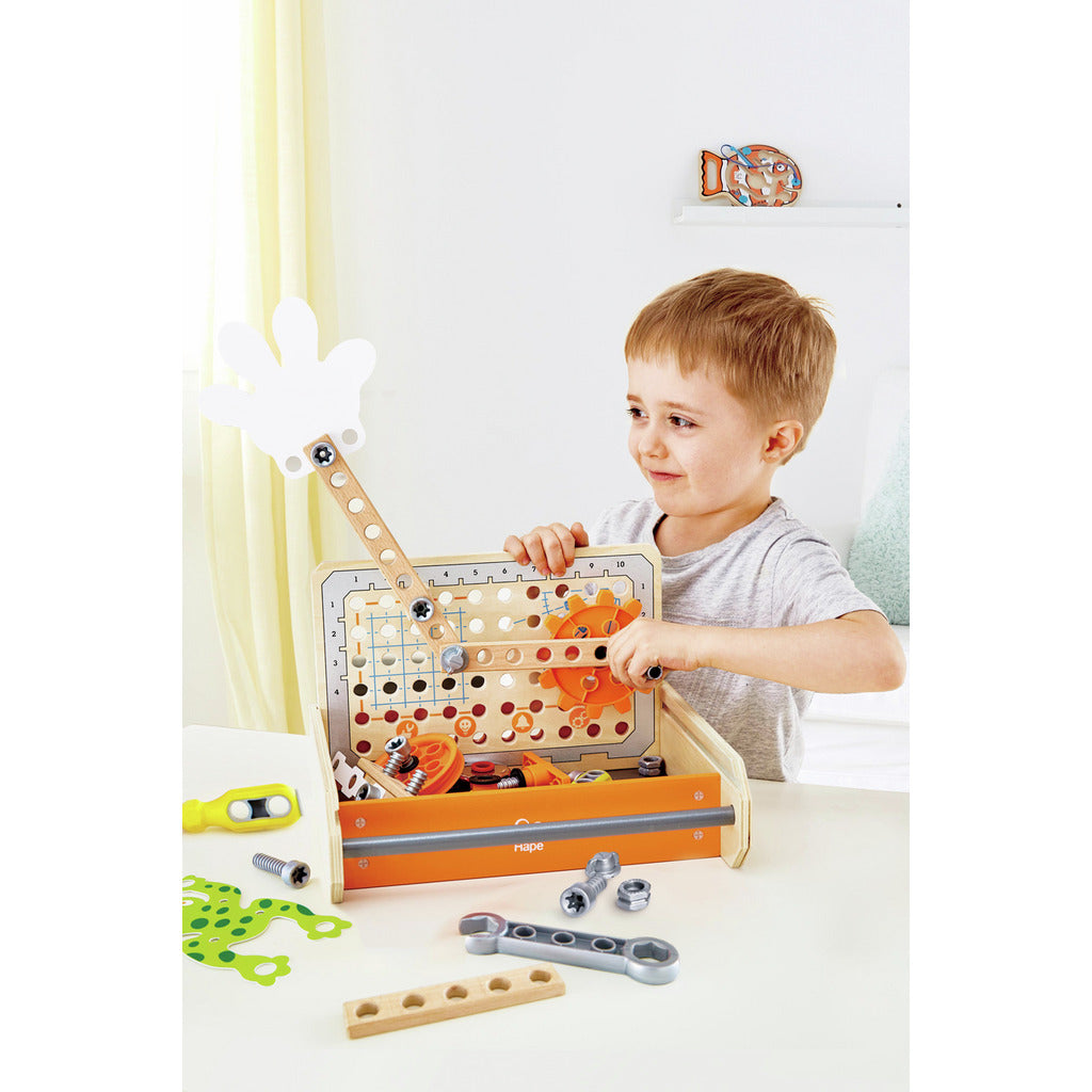 Smiling boy plays with a wooden science experiment toolbox assembling orange gears and manipulating a white hand lever with loose tools nearby