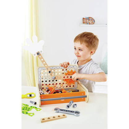 Smiling boy plays with a wooden science experiment toolbox assembling orange gears and manipulating a white hand lever with loose tools nearby
