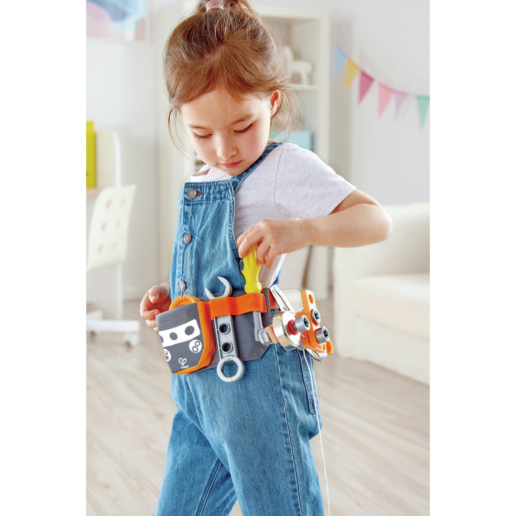 Young girl wearing denim overalls interacts with the orange Scientific Tool Belt, holding a yellow screwdriver while standing in a playroom