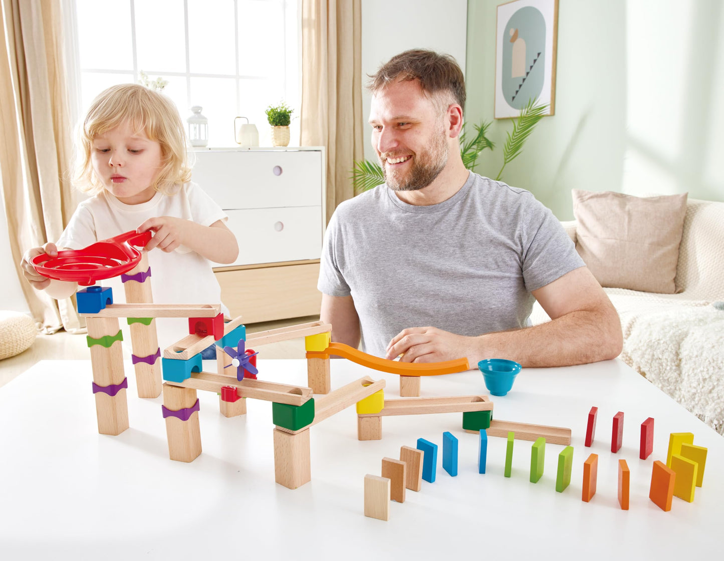 A child drops a marble into the red funnel of a wooden marble run and domino track while an adult smiles next to them