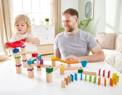 A child drops a marble into the red funnel of a wooden marble run and domino track while an adult smiles next to them