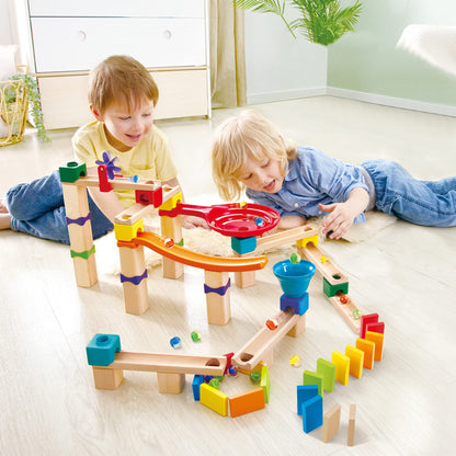 Two enthusiastic boys build and interact with a tall wooden marble run featuring an orange ramp, red spiral track, blue funnel, purple pinwheel, and multicolored dominoes