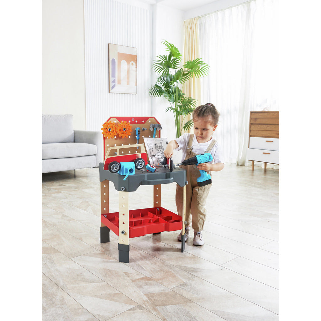 A child stands at the red and gray toy vehicle service bench, which features a van motif, large gears, and holds a blue toy power drill