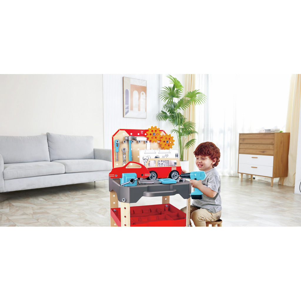 A young boy uses a blue toy drill while sitting at the wooden Vehicle Service and Repair Workbench which features a red car and orange gears