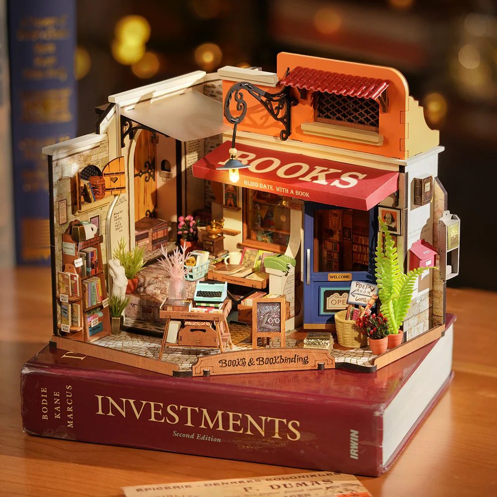 Intricate DIY wooden miniature corner bookstore model with a red awning, tiny books, and a working light, displayed resting on a large burgundy textbook