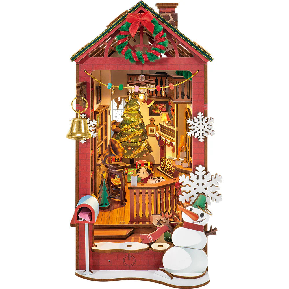 Miniature DIY Christmas bookend kit showing a festive red house diorama with a brightly lit tree, snowman, wreath, and golden bell