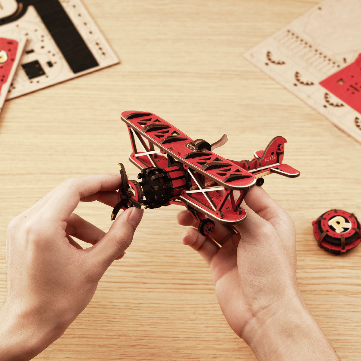 Two hands hold the finished mechanical red wooden biplane model from the Rokr Sky Captain kit above laser-cut assembly boards