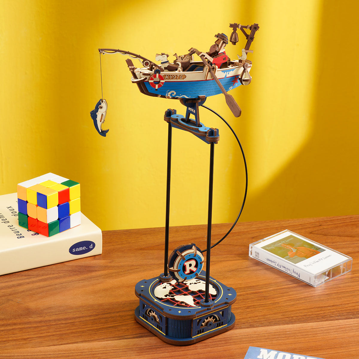 An intricate ROKR wooden model features a blue fishing boat with a fisherman and a dog suspended over a detailed dark blue base showing a world map