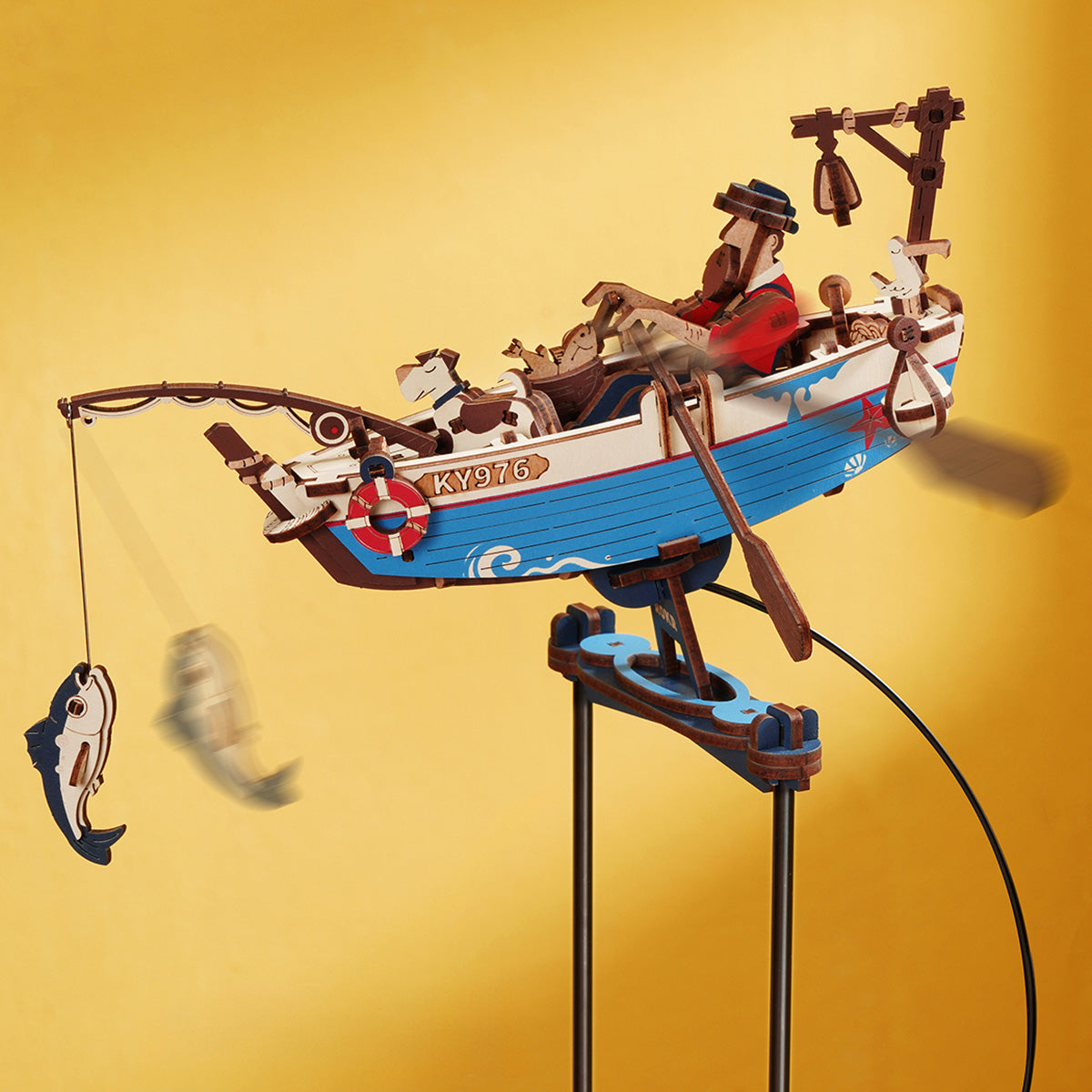 The Rokr Gravity Swing Ocean Fisher 3D wooden puzzle features a blue fishing boat and a fisherman figure with a dog mounted on a stand while a fish swings below
