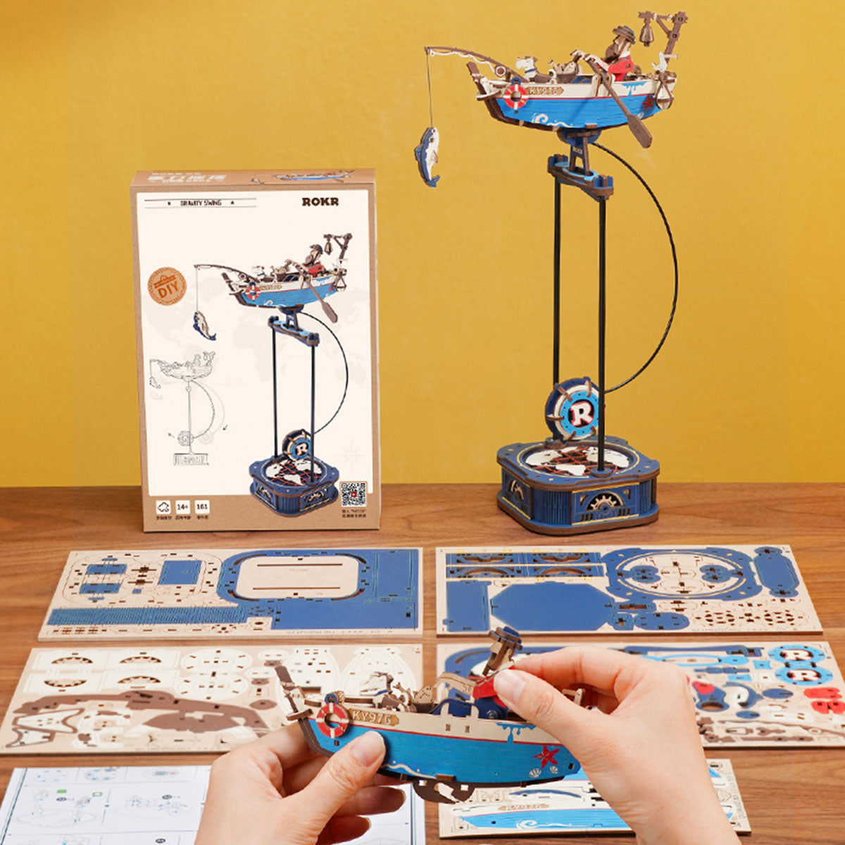 Hands assemble the detailed blue and wood ROKR Gravity Swing Ocean Fisher boat model displayed next to its box and the finished kinetic sculpture