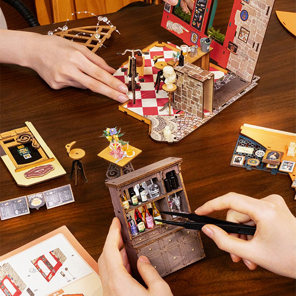 People meticulously build the miniature Herys Chocolate cafe diorama, placing tiny bottles into a detailed wooden cabinet using precision tweezers