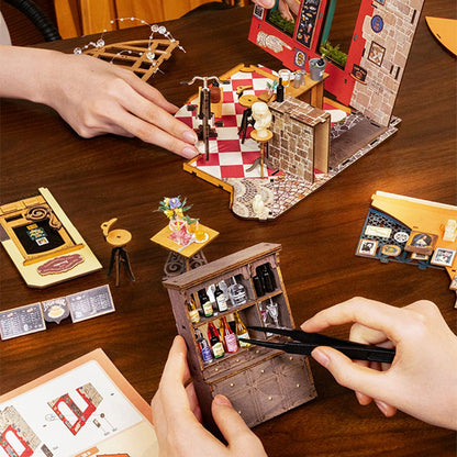 People meticulously build the miniature Herys Chocolate cafe diorama, placing tiny bottles into a detailed wooden cabinet using precision tweezers