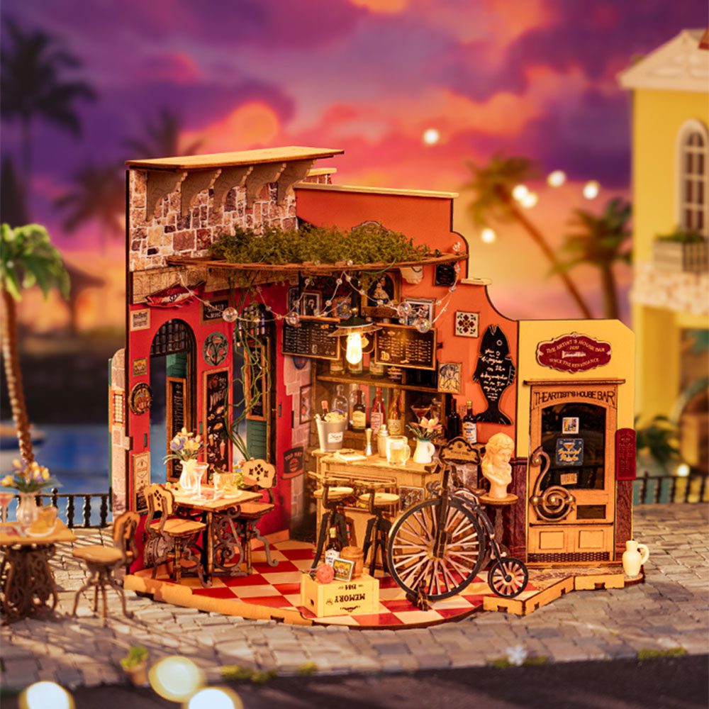 Miniature wooden DIY model of an illuminated street bar and cafe, showing checkered tile floors, detailed furnishings, and a tropical sunset backdrop