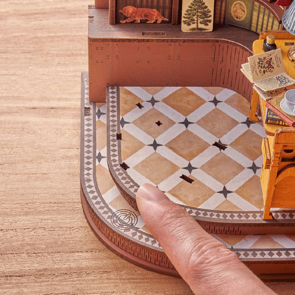 A detailed view of the Silent Corner Study DIY bookend kit shows a human finger emphasizing the tiny scale of the curved, patterned miniature floor and stairs