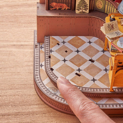 A detailed view of the Silent Corner Study DIY bookend kit shows a human finger emphasizing the tiny scale of the curved, patterned miniature floor and stairs