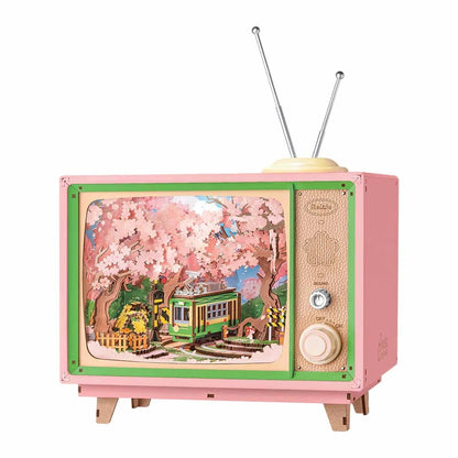 Pink wooden music box shaped like a retro TV with antennae, showing a miniature diorama of a green tram passing through dense pink cherry blossoms
