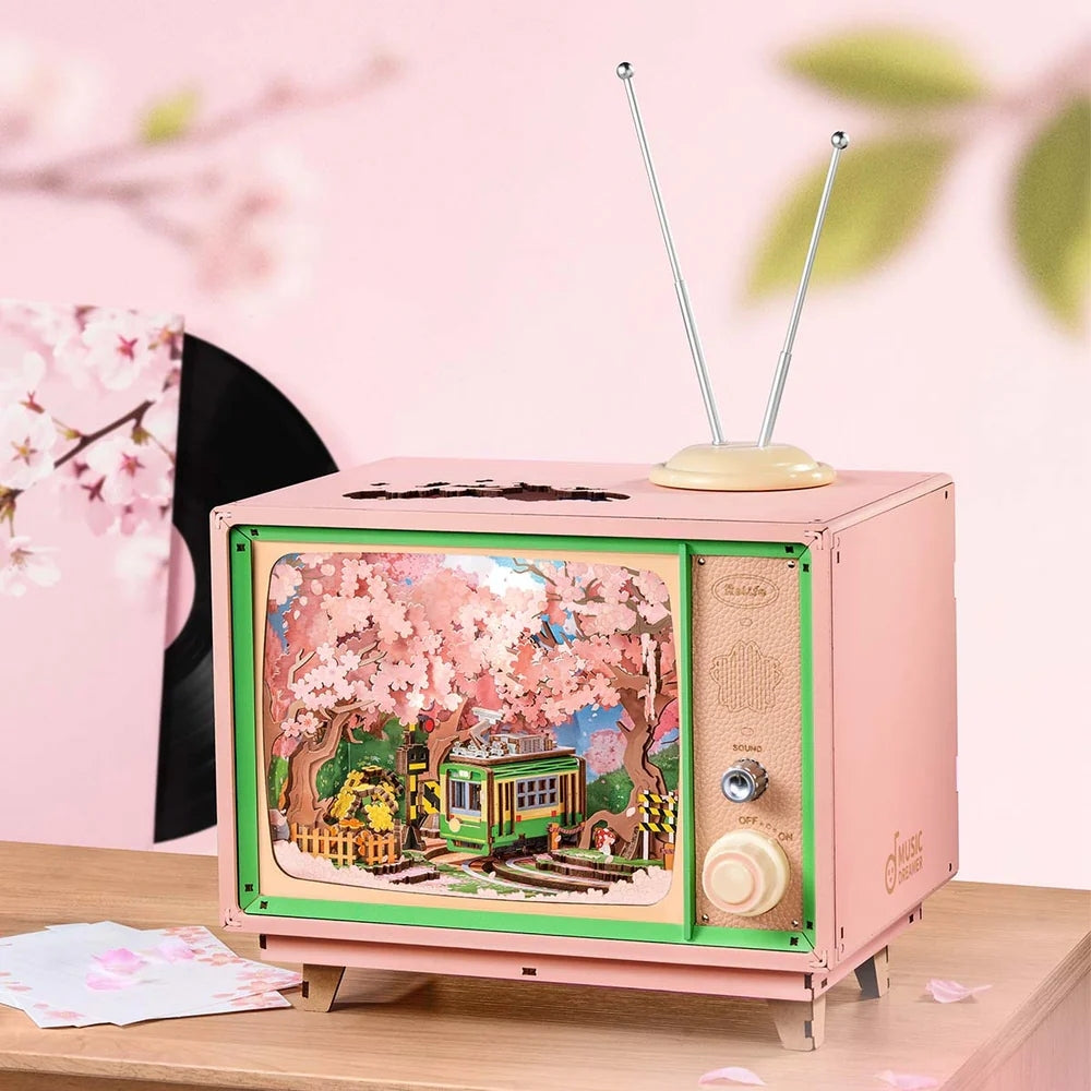 This charming pink retro television music box holds a detailed miniature diorama showing a green trolley moving through a vibrant grove of cherry blossom trees