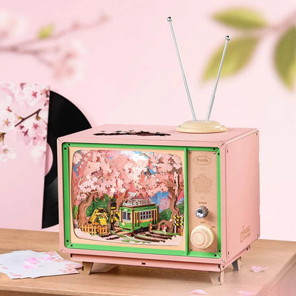 This charming pink retro television music box holds a detailed miniature diorama showing a green trolley moving through a vibrant grove of cherry blossom trees
