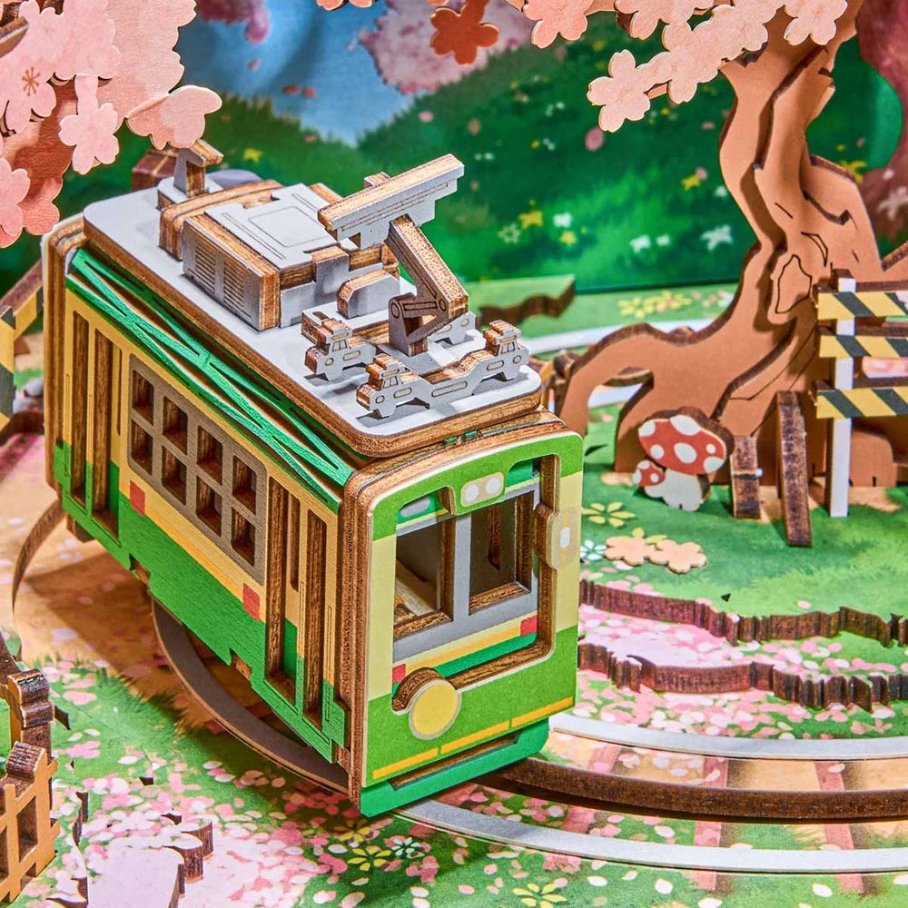 The assembled wooden model of a bright green and beige streetcar kit set on miniature tracks surrounded by decorative pink sakura foliage and grass