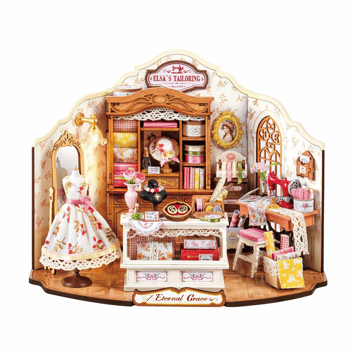 This DIY miniature house scene captures Elsa's Tailoring shop showing a tiny sewing machine, rolls of fabric, a floral dress on a mannequin, and antique wooden shelving