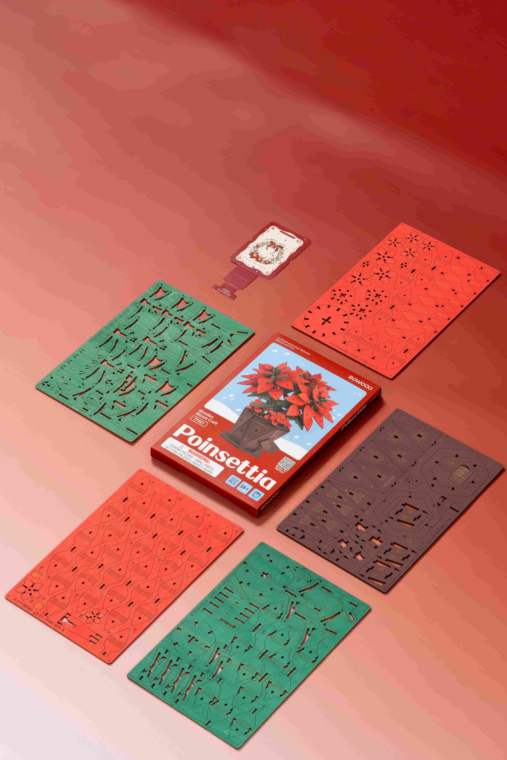 The Rowood Poinsettia Wooden Bloom Craft kit box, showing a completed red flower, is displayed with multiple component sheets in corresponding shades of red, green, and dark brown