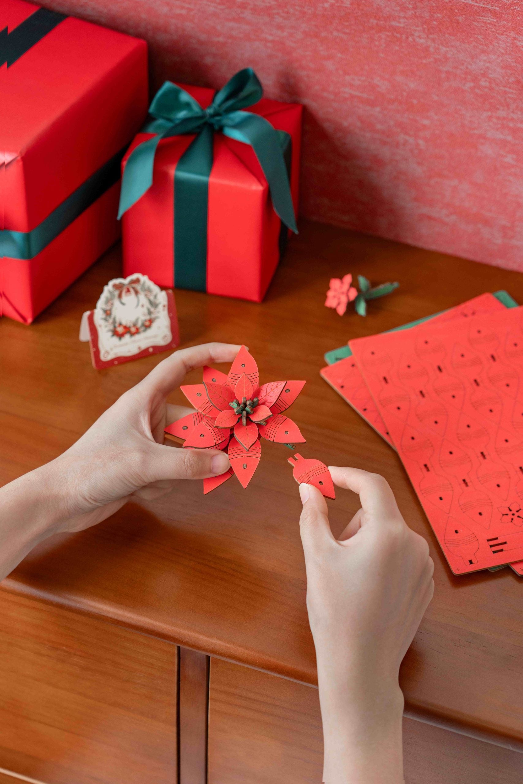 The Wood Bloom Poinsettia puzzle is being assembled by hand, featuring bright red die-cut wood pieces and a festive holiday background