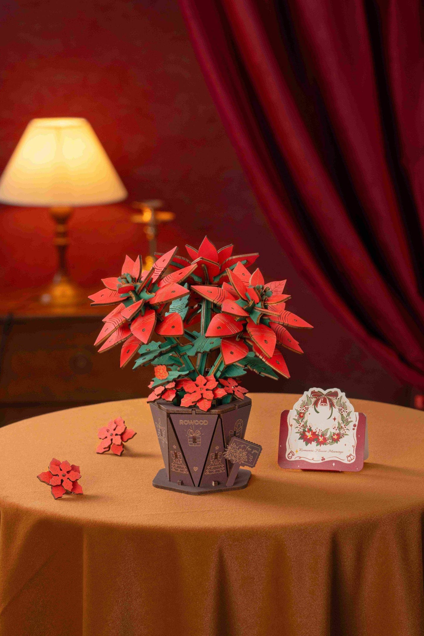 The vibrant red and green 3D wooden poinsettia model is displayed in a small hexagon-shaped pot next to two loose petals and an elegant holiday card