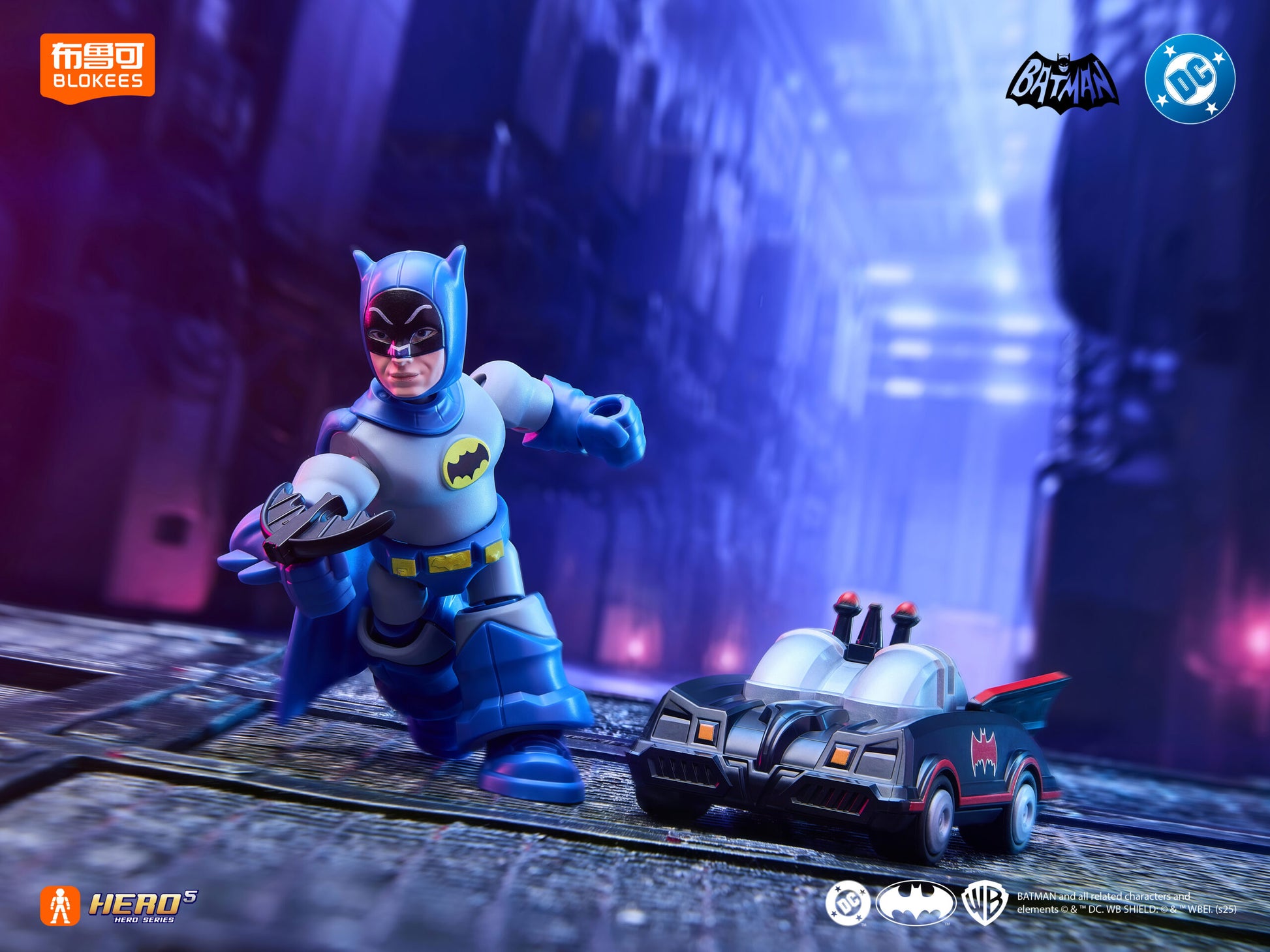 Blue and gray Batman action figure holding a weapon, standing beside a small Batmobile in a neon-lit city