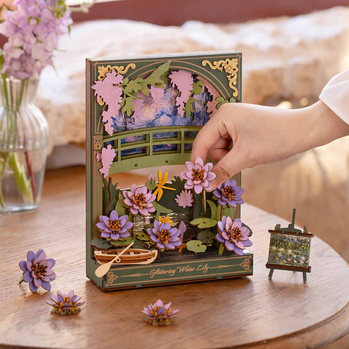 Hand placing a purple wooden water lily onto a 3D craft puzzle depicting a pond and green bridge