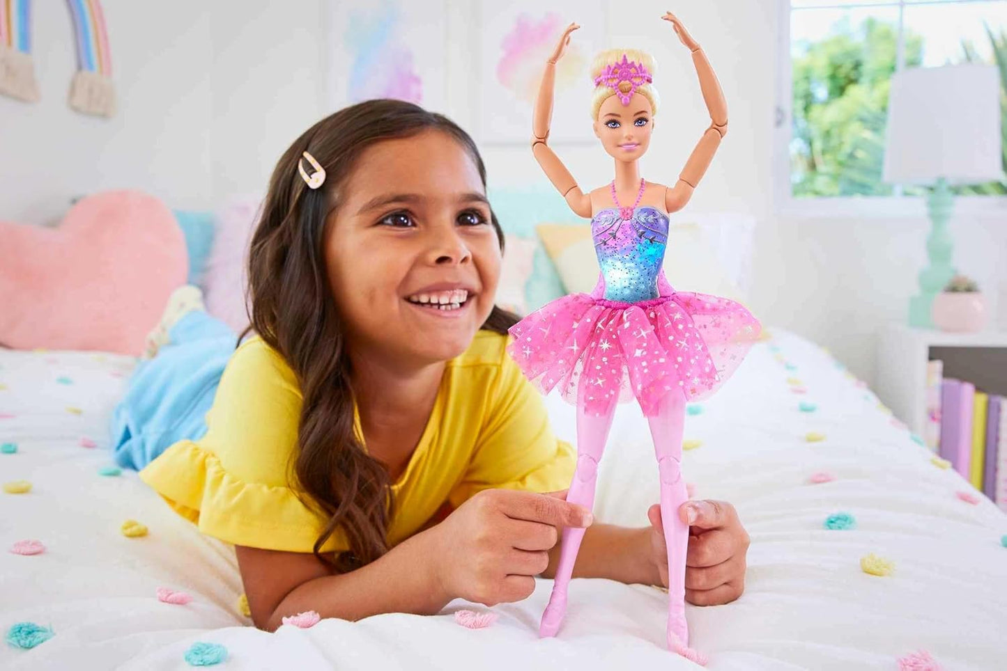 A smiling girl on a colorful bed holds a ballerina Barbie doll with raised arms -  wearing a pink tutu and tiara