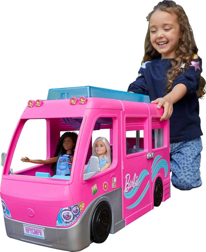 A smiling girl plays with a pink Barbie camper van that has two Barbie dolls seated inside