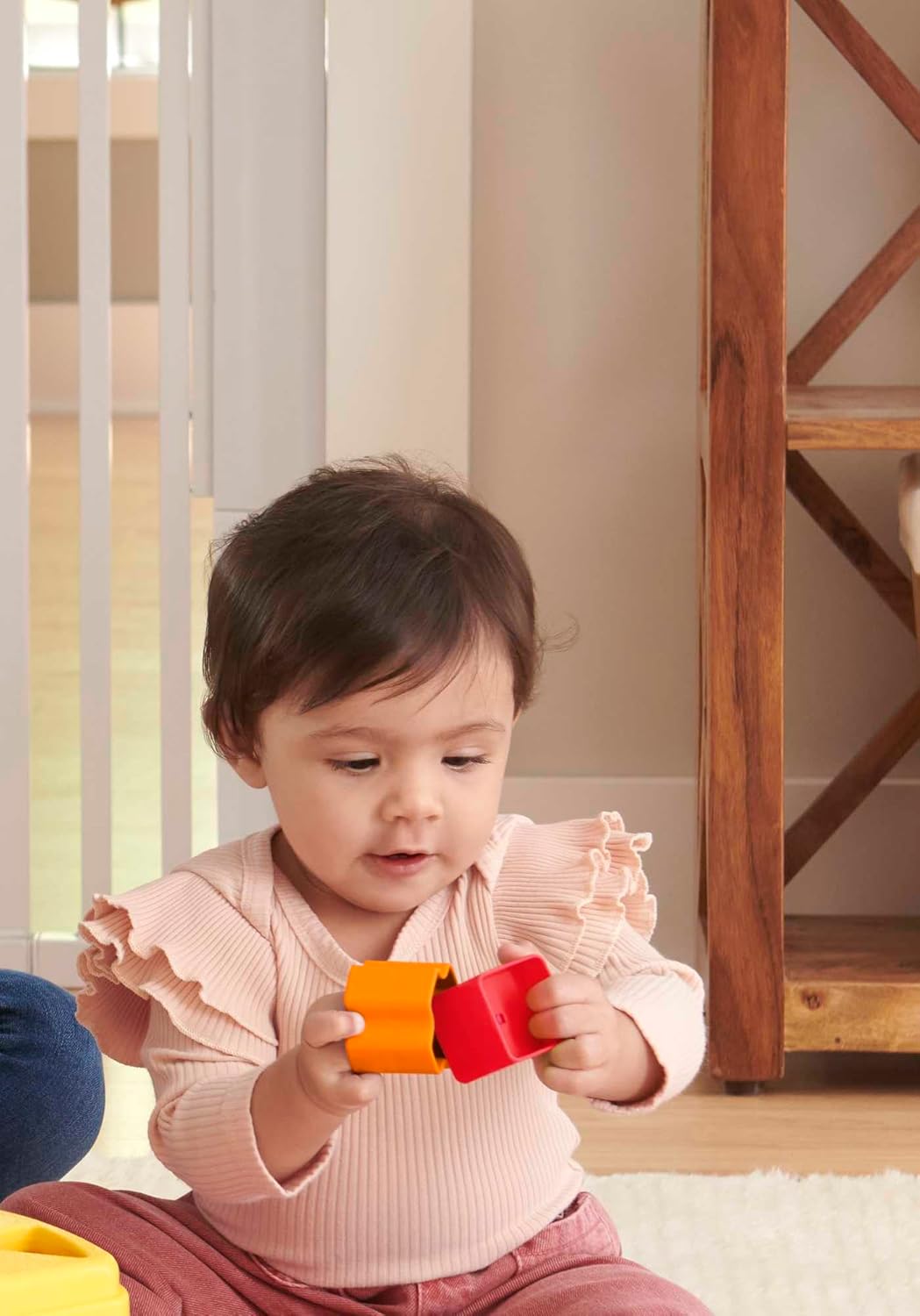 A baby with dark hair intently connects orange and red toy blocks while sitting on a rug