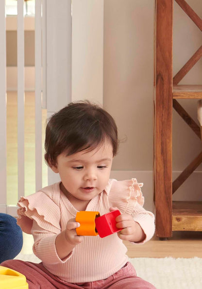 A baby with dark hair intently connects orange and red toy blocks while sitting on a rug