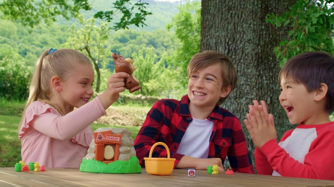 Three joyful children play an outdoor Beware of The Bear board game, a girl holds a toy bear while two boys laugh and clap