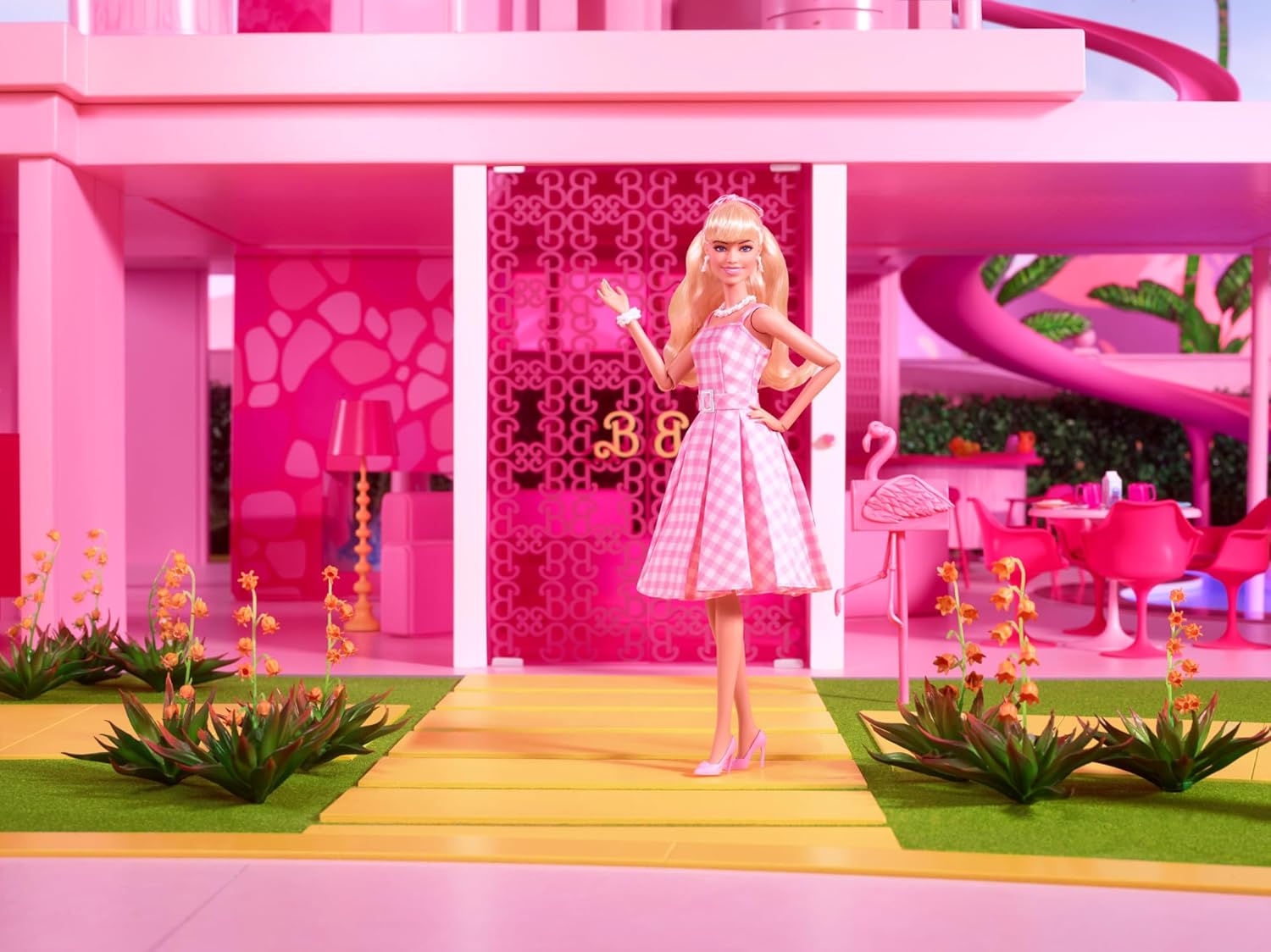 Blonde Barbie in pink gingham dress smiles -  waving from her iconic all-pink Dreamhouse with a yellow path