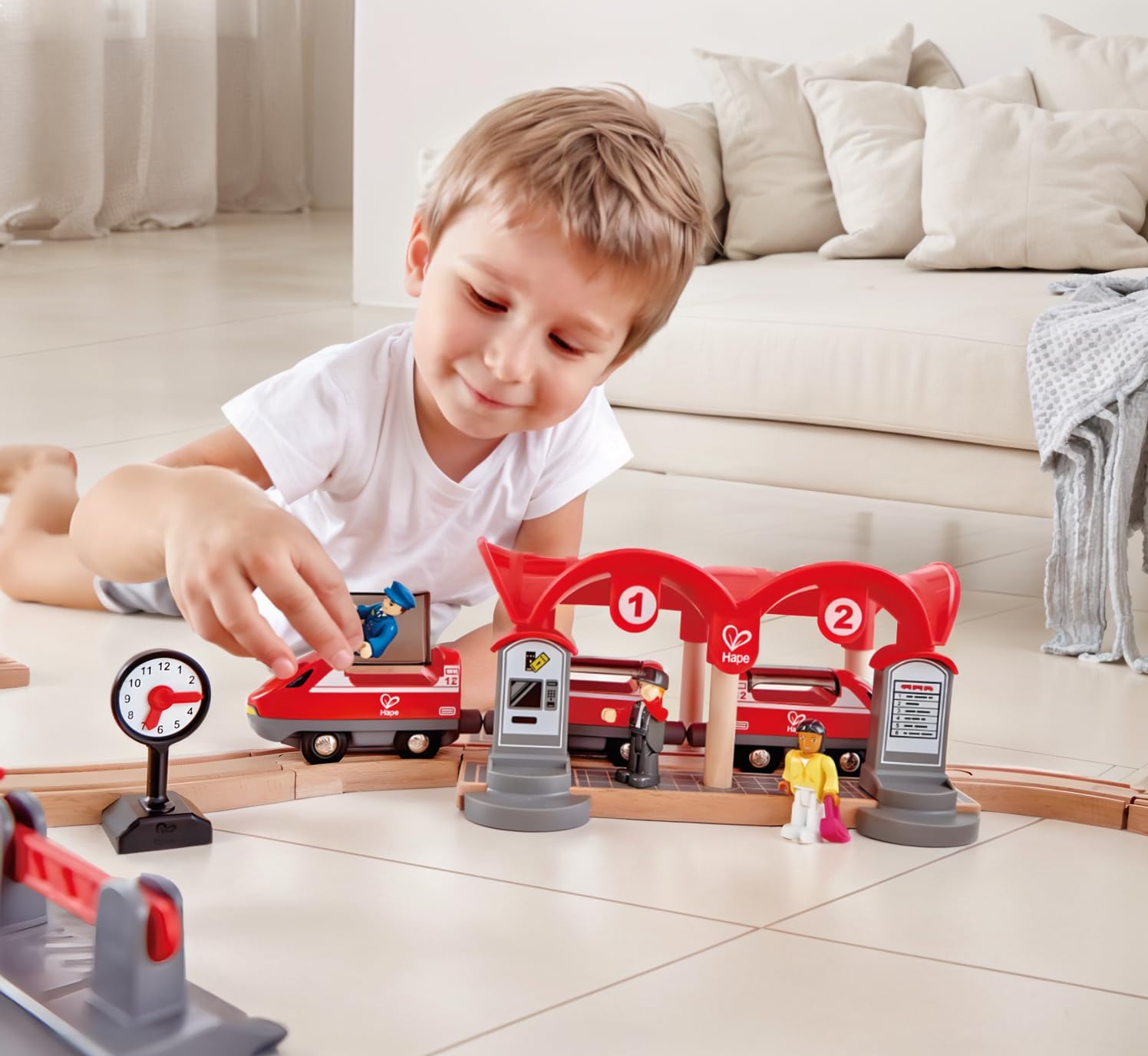 Smiling boy plays on the floor, placing a figure onto a red toy train at a wooden train station set