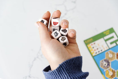 A hand grasps several white game dice with various resource symbols, set against a blurred board game