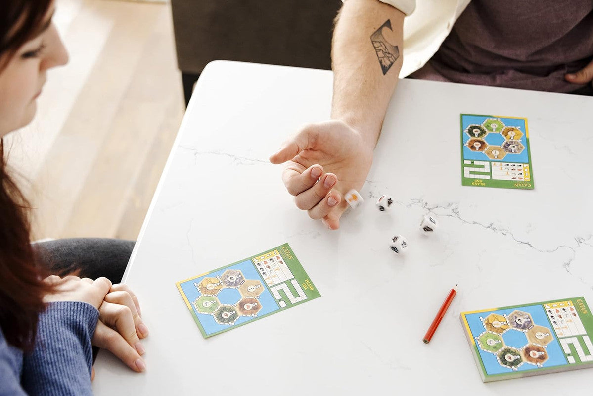 A man's hand rolls dice on a white table with Catan scorecards, while a woman watches