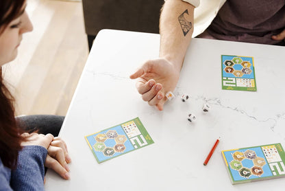 A man's hand rolls dice on a white table with Catan scorecards, while a woman watches