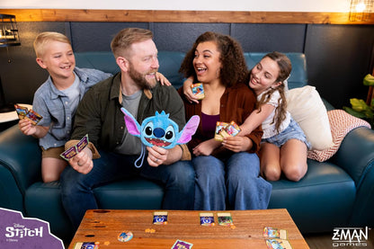 Four people on a couch playing the Disney Stitch card game, laughing, with a Stitch plush
