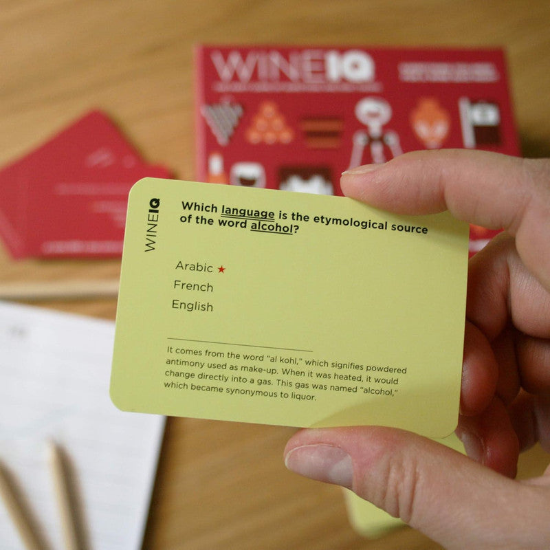 Hand holds a yellow Wine IQ trivia card asking Which language is the etymological source of the word alcohol