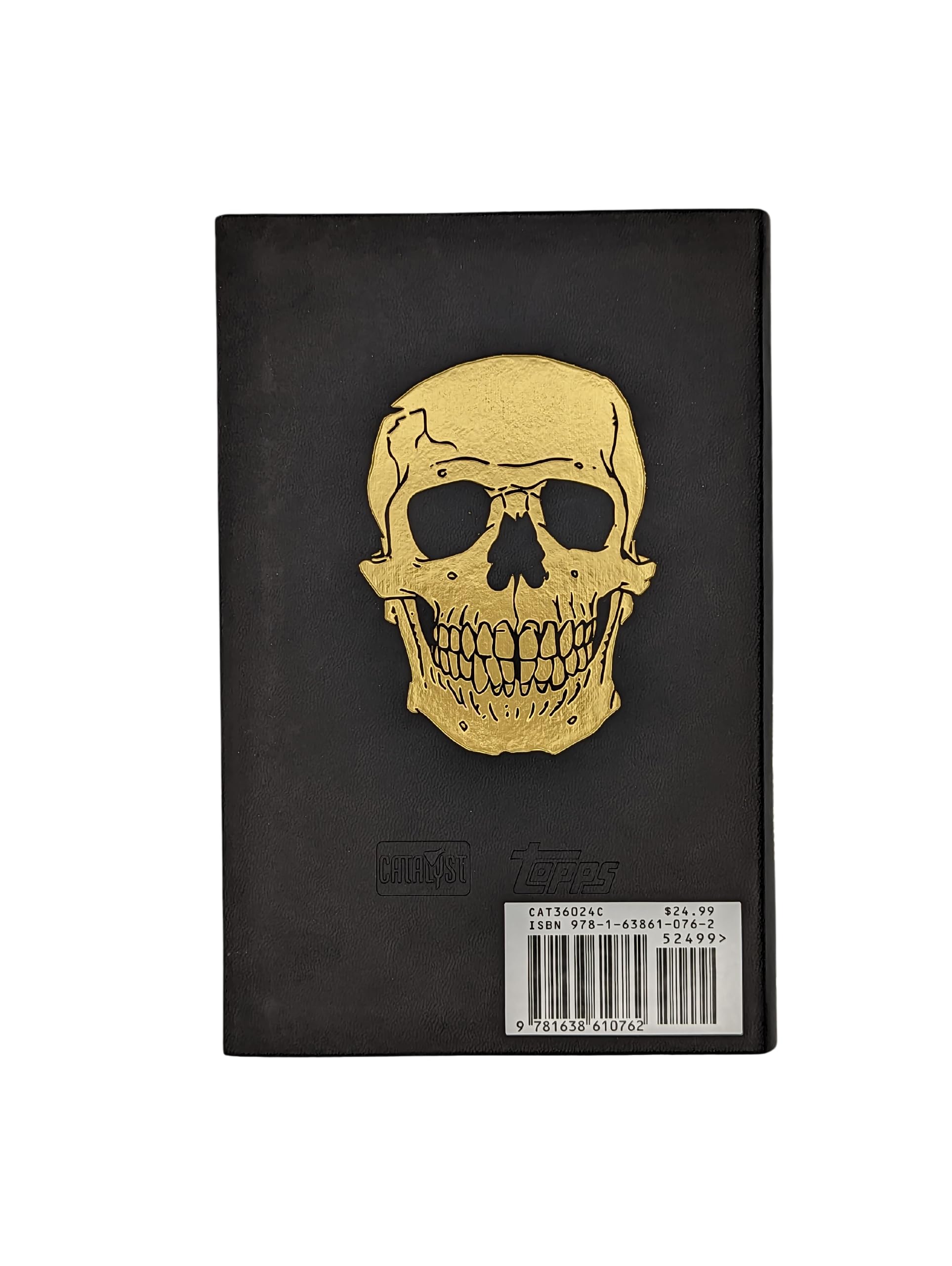 Black book with a prominent golden skull on the front cover. Topps & Catalyst logos, barcode, and price are at the bottom