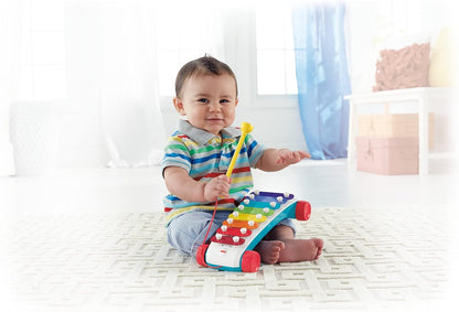 A smiling baby on a rug holds a yellow mallet above a colorful toy xylophone