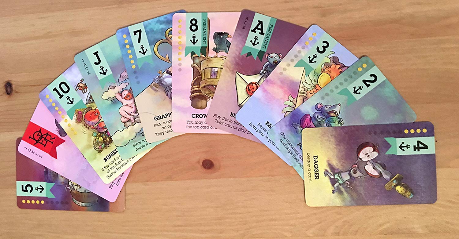 A fan of colourful Scuttle! The Curse of Black Jack Expansion cards, featuring various fantasy illustrations, numbers, and anchor symbols, is spread on a wooden table