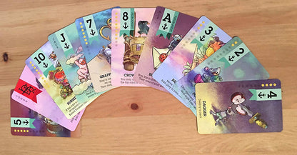 A fan of colourful Scuttle! The Curse of Black Jack Expansion cards, featuring various fantasy illustrations, numbers, and anchor symbols, is spread on a wooden table