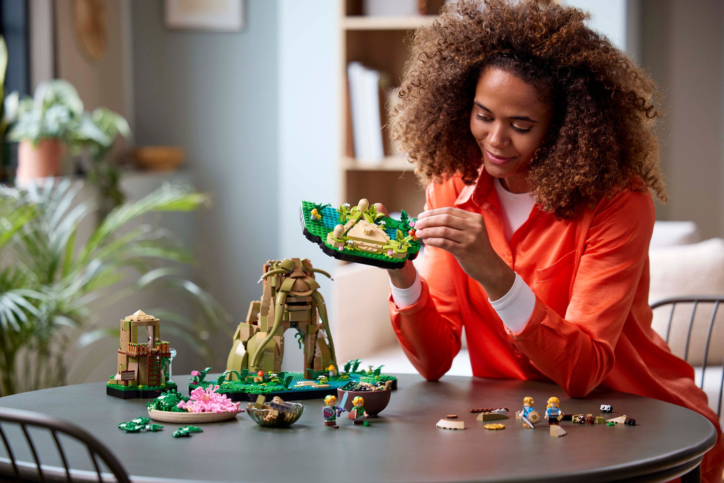 A smiling woman in an orange shirt builds a detailed brick model of a fantasy landscape on a dark table