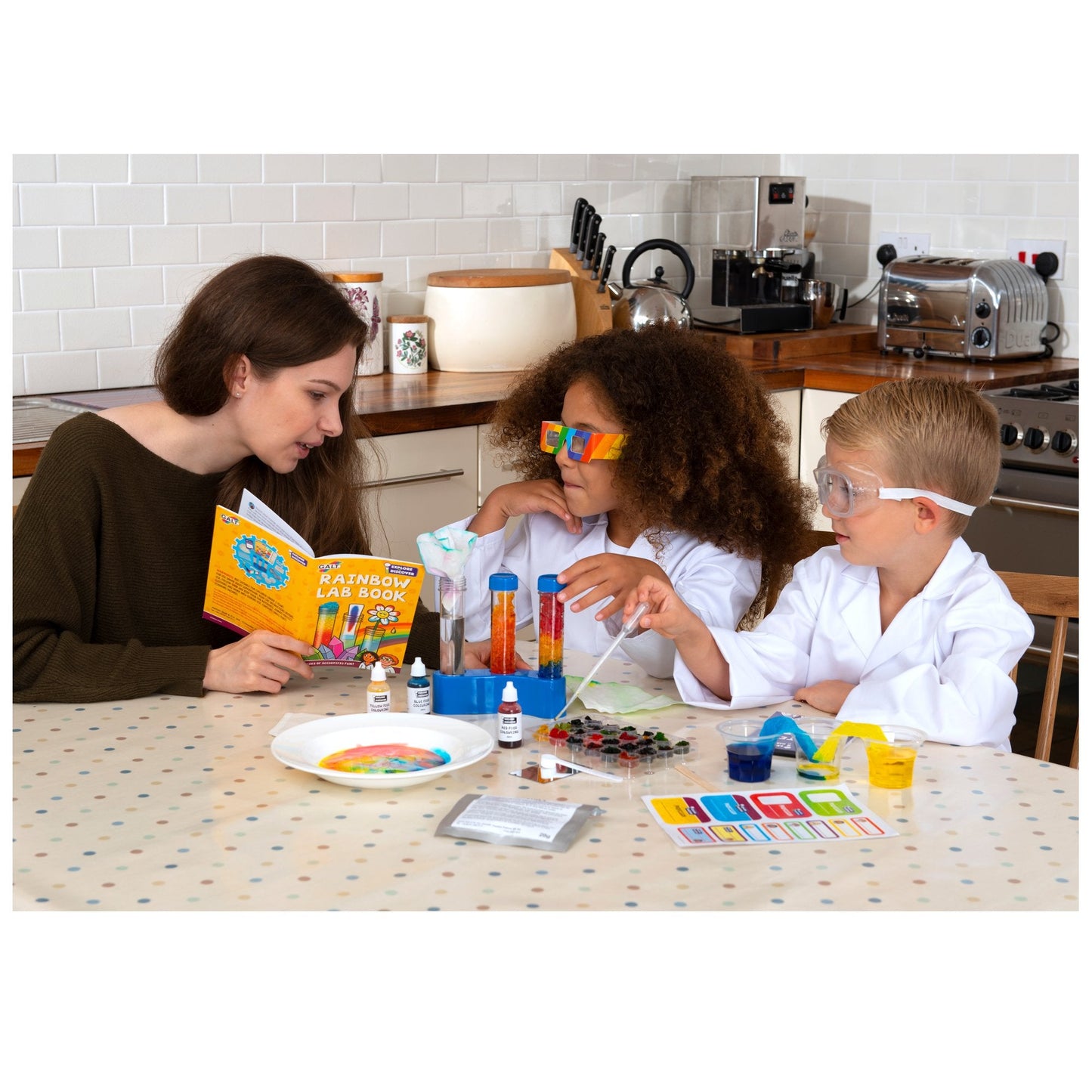 Adult reads from a "Rainbow Lab Book" instructing two children in lab coats performing a colourful science experiment