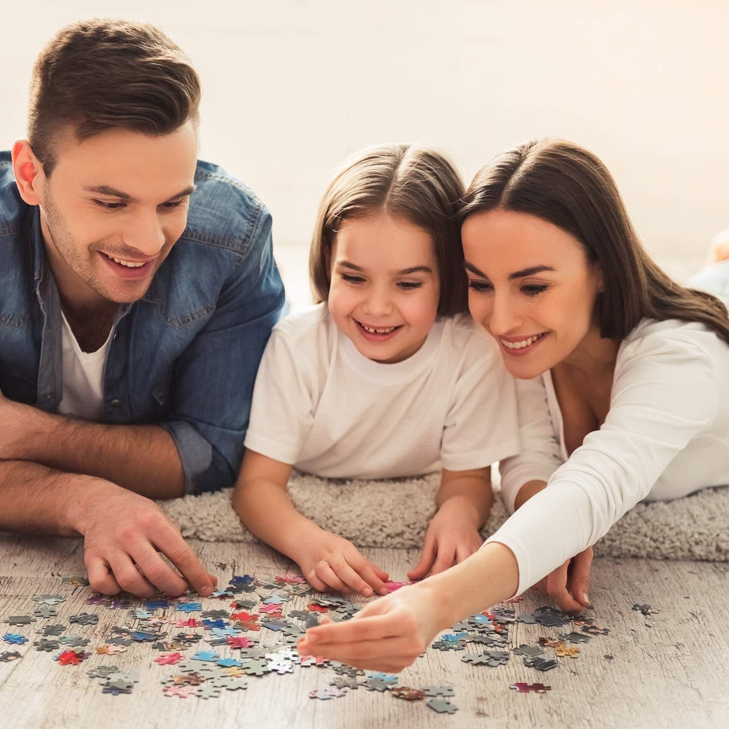 A happy family featuring a smiling mother, father, and young daughter lies on the floor collaborating on the colorful 1000 piece jigsaw puzzle