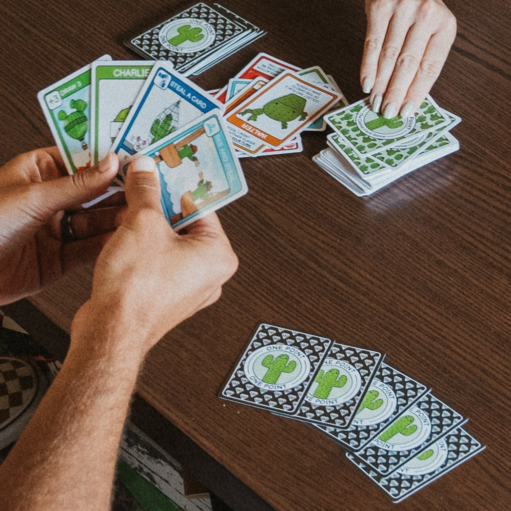 Two people play the Cactus Card Game, dealing green-backed cards and holding up several illustrated character and action cards featuring cartoon cacti